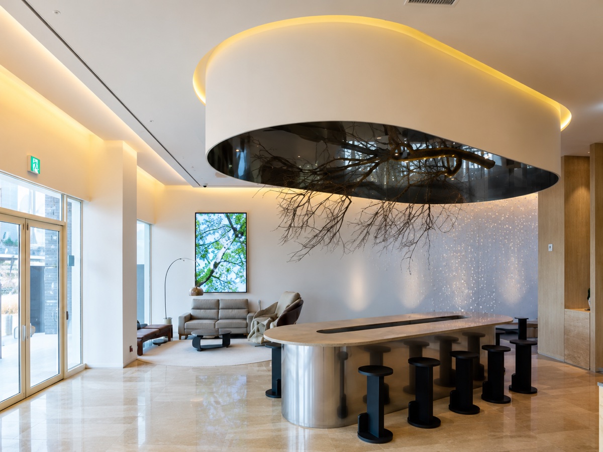 The interior of a premium learning centre: a long stone table ringed with stools under a backlit oval ceiling feature, with lounge seating beside full-height windows.