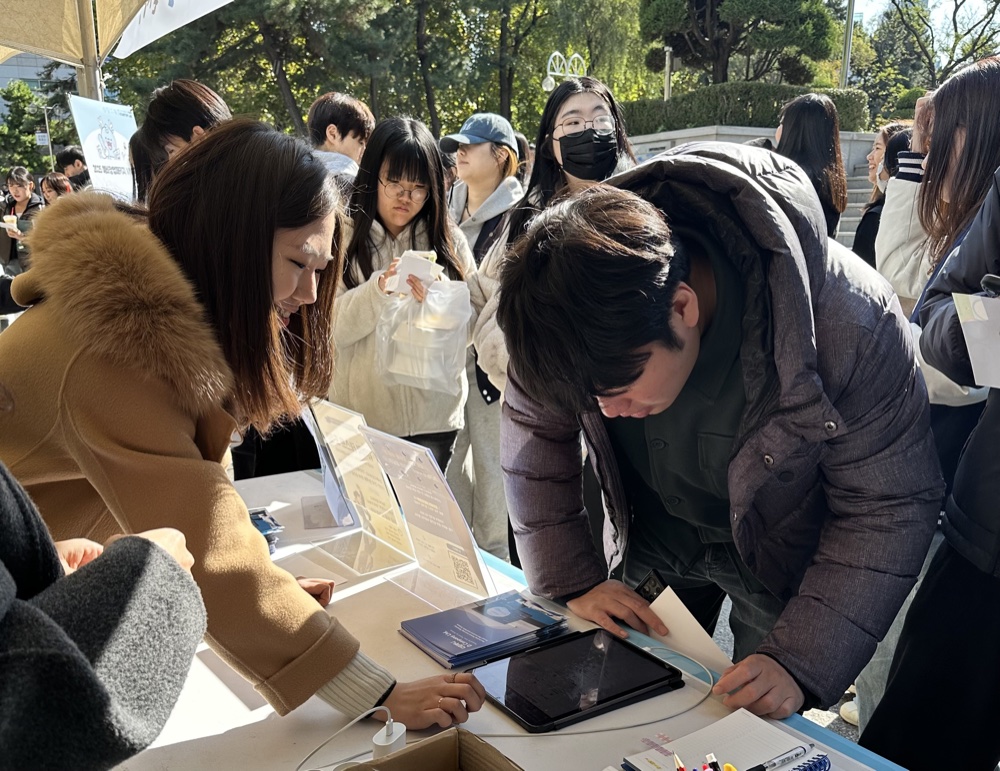 University students gathered around an outdoor D.Company booth on campus, trying the L:BRN app on a tablet while staff members guide them through the demo.